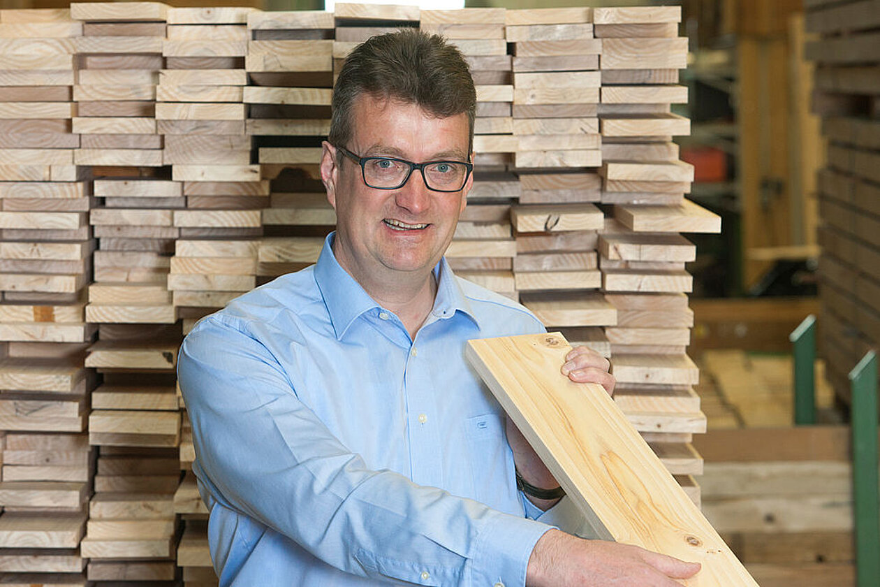 Fachgerechte Beratung rund um die Gutshofdiele vom Fachhändler Mann in blauem Hemd, hält eine Holzdiele in der Hand; im Hintergrund gestapelte Holzbohlen